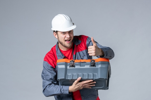 Vista frontal del constructor masculino en uniforme tratando de abrir la caja de herramientas sobre fondo gris