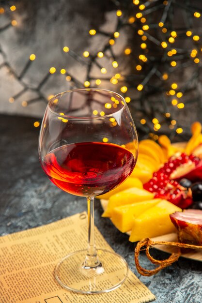 Vista frontal copa de vino uvas piezas de queso rebanadas de carne en un periódico de placa de madera en luces de Navidad oscuras
