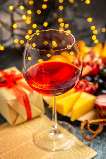 Vista frontal copa de vino uvas piezas de queso rebanadas de carne en placa de madera en luces de Navidad oscuras