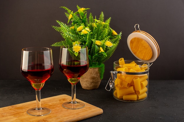 Foto gratuita una vista frontal copas de vino en un escritorio de madera marrón junto con flores y pasta italiana cruda en un escritorio oscuro