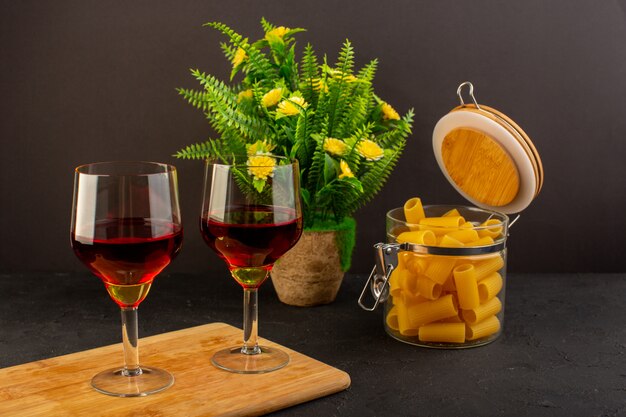 Una vista frontal copas de vino en un escritorio de madera marrón junto con flores y pasta italiana cruda en un escritorio oscuro