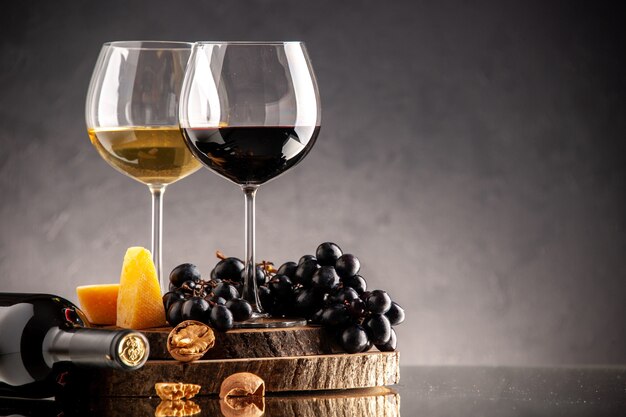 Vista frontal copas de vino uvas frescas nueces queso amarillo sobre tablero de madera botella volcada sobre fondo oscuro
