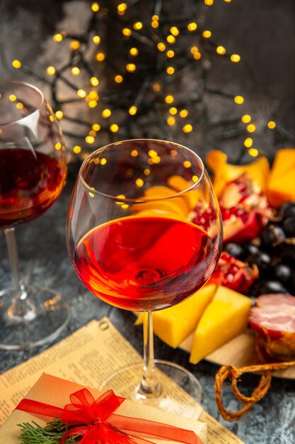 Vista frontal copas de vino uvas piezas de queso rebanadas de carne en placa de madera en luces de Navidad oscuras