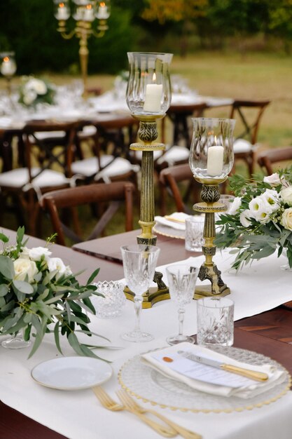 Vista frontal de cristalería y cubertería servida en la mesa de madera con composiciones florales y candelabros al aire libre