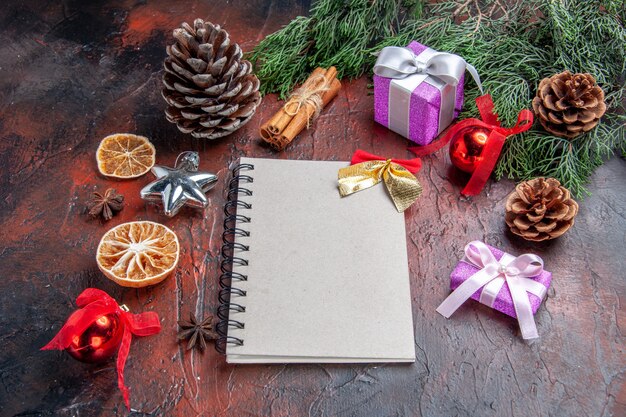 Vista frontal de un cuaderno con pequeño arco ramas de pino conos árbol de navidad juguetes y regalos anís estrellado de canela sobre fondo rojo oscuro