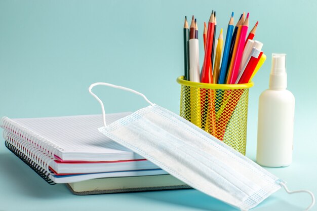Vista frontal de cuadernos y bolígrafos, lápices de colores, máscara y spray sobre superficie azul