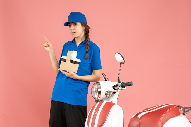 Vista frontal de la curiosa chica de mensajería de pie junto a la motocicleta sosteniendo café apuntando hacia arriba sobre fondo de color melocotón pastel
