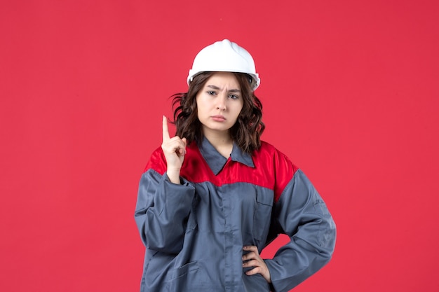 Vista frontal de la curiosa constructora en uniforme con casco y apuntando hacia arriba sobre fondo rojo aislado