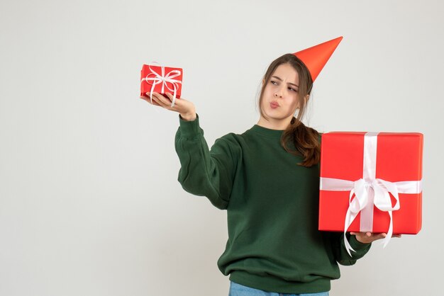 Vista frontal curiosa niña con gorro de fiesta comparando sus regalos de Navidad