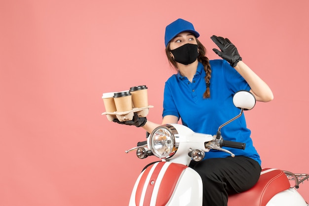 Vista frontal de la curiosa repartidora femenina con máscara médica y guantes sentado en scooter con órdenes sobre fondo de melocotón pastel