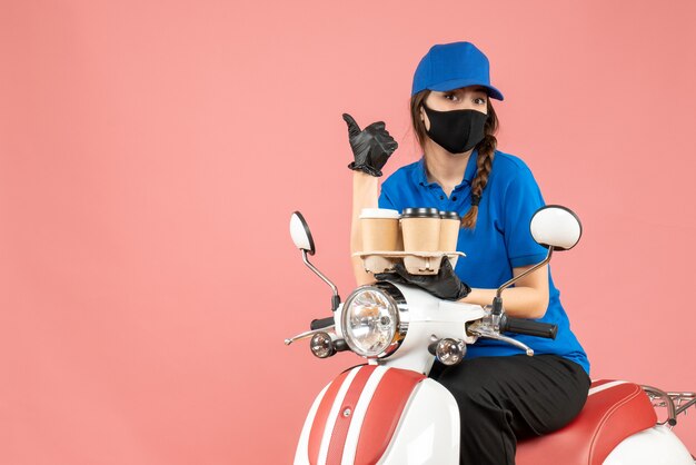 Vista frontal de la curiosa repartidora con máscara médica y guantes sentado en scooter sosteniendo pedidos apuntando hacia arriba sobre fondo melocotón pastel