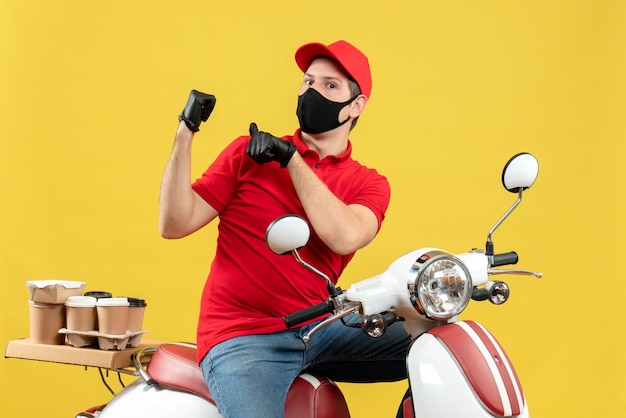 Vista frontal del curioso adulto joven vistiendo blusa roja y guantes de sombrero en máscara médica entregando orden sentado en scooter apuntando hacia atrás sobre fondo amarillo