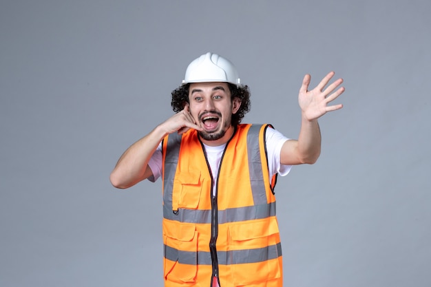 Foto gratuita vista frontal del curioso constructor masculino vistiendo chaleco de advertencia con casco de seguridad y haciendo gesto de llamarme en la pared de onda gris