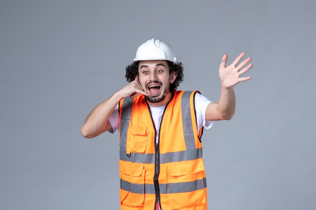 Vista frontal del curioso constructor masculino vistiendo chaleco de advertencia con casco de seguridad y haciendo gesto de llamarme en la pared de onda gris