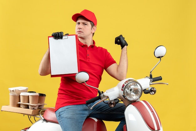 Foto gratuita vista frontal del curioso mensajero hombre vestido con blusa roja y guantes de sombrero en máscara médica entregando orden sentado en scooter sosteniendo documento