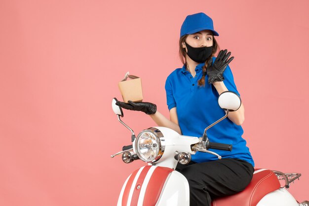 Vista frontal del curioso repartidor con máscara médica y guantes sentado en scooter entregando pedidos sobre fondo melocotón pastel