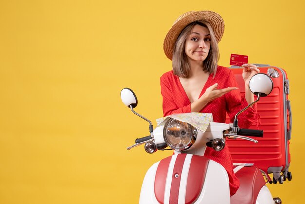 Vista frontal de la dama rubia en vestido rojo con tarjeta de descuento cerca de ciclomotor