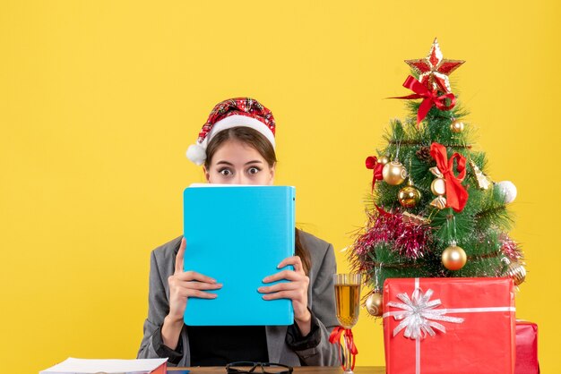 Foto gratuita vista frontal deambulaba niña con sombrero de navidad sentado en la mesa cubriendo la cara con carpeta árbol de navidad y cóctel de regalos