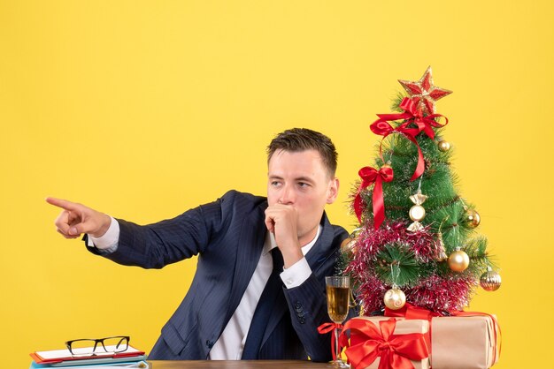 Vista frontal del dedo del hombre curioso señalando algo sentado en la mesa cerca del árbol de Navidad y regalos en amarillo