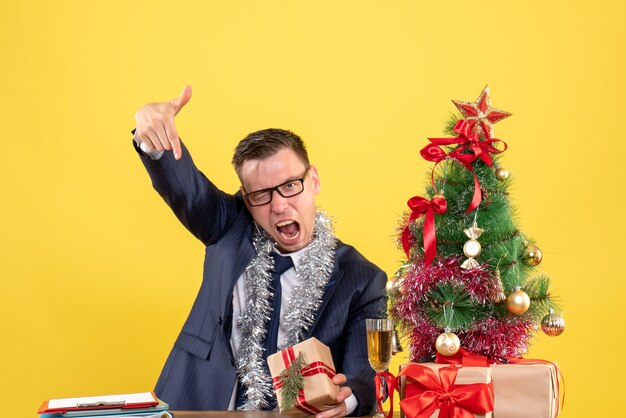 Vista frontal del dedo del hombre enojado apuntando hacia abajo sentado en la mesa cerca del árbol de Navidad y presenta en la pared amarilla