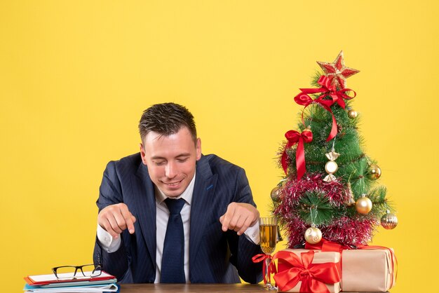 Vista frontal del dedo del hombre sonriente apuntando a la mesa sentado en la mesa cerca del árbol de Navidad y regalos en amarillo