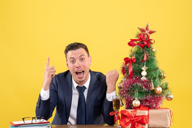 Vista frontal del dedo del hombre sorprendido apuntando hacia arriba sentado en la mesa cerca del árbol de Navidad y regalos en la pared amarilla