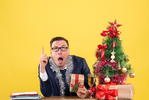 Vista frontal del dedo del hombre sorprendido apuntando hacia arriba sentado en la mesa cerca del árbol de Navidad y regalos en la pared amarilla