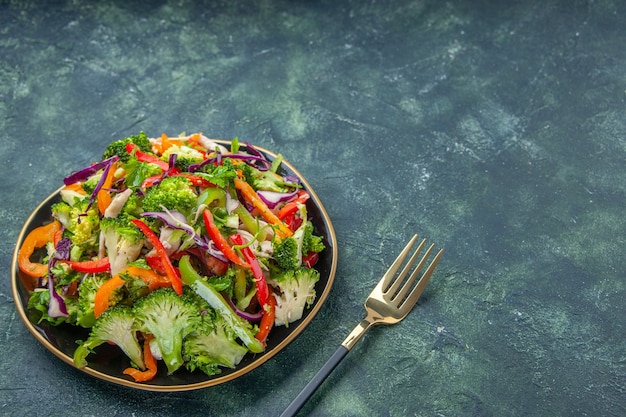 Vista frontal de una deliciosa ensalada vegana en un plato con varias verduras y un tenedor en el lado derecho sobre un fondo oscuro con espacio libre