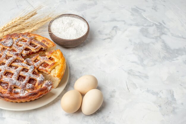 Vista frontal de la deliciosa tarta de mermelada en un plato blanco, huevos con picos de harina en una olla pequeña sobre una mesa blanca con espacio libre