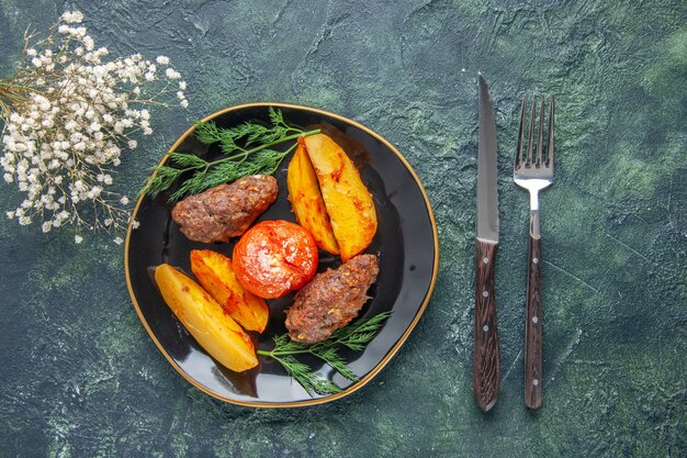 Vista frontal de deliciosas chuletas de carne al horno con patatas y tomates en un plato negro cubiertos con flores blancas sobre fondo verde negro de colores mezclados