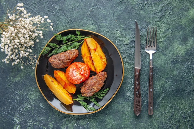 Vista frontal de deliciosas chuletas de carne al horno con patatas y tomates en un plato negro cubiertos con flores blancas sobre fondo verde negro de colores mezclados