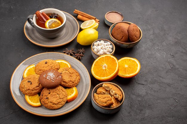 Vista frontal deliciosas galletas de arena con naranjas frescas y una taza de té sobre fondo oscuro galleta de fruta galleta dulce azúcar cítrico