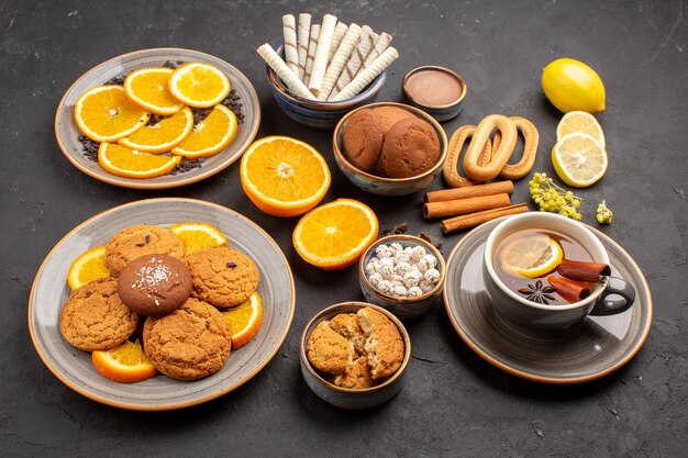 Vista frontal deliciosas galletas de azúcar con una taza de té y naranjas sobre fondo oscuro azúcar té fruta galleta galleta dulce