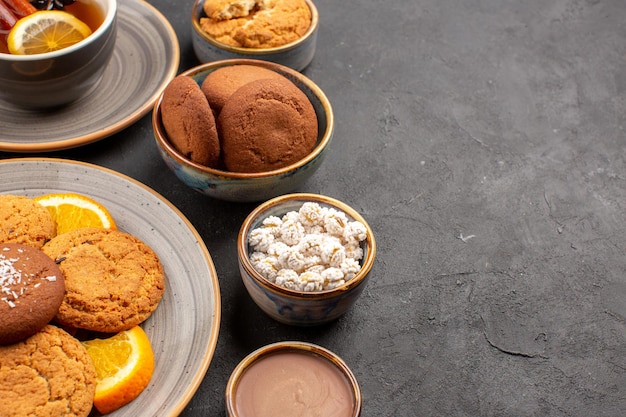 Vista frontal deliciosas galletas con una taza de té y naranjas en rodajas sobre fondo oscuro galleta fruta dulce pastel cookie cítricos