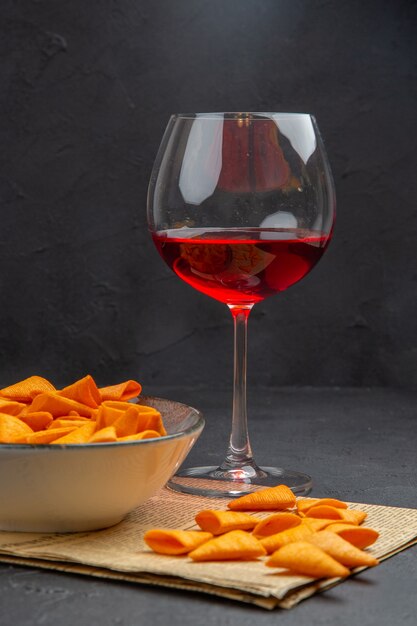 Vista frontal de deliciosas papas fritas dentro y fuera del recipiente y vino tinto en un vaso en un periódico viejo