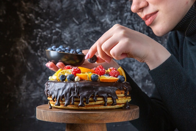 Vista frontal delicioso pastel de chocolate decorado con frutas por mujer en pared oscura