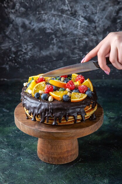 Vista frontal delicioso pastel de chocolate con frutas cortadas por mujer en la pared oscura