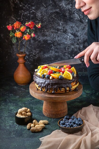 Vista frontal delicioso pastel de chocolate con frutas cortadas por mujer en la pared oscura