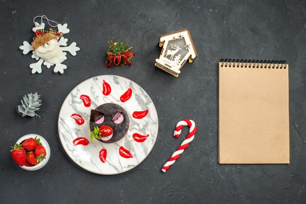 Vista frontal delicioso pastel de queso con fresa y chocolate en plato ovalado cuenco de fresas cuaderno de juguetes de árbol de Navidad sobre fondo oscuro