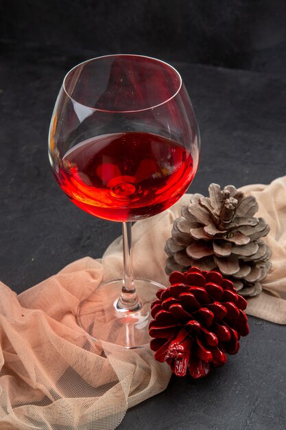 Vista frontal del delicioso vino tinto en una copa de cristal sobre una toalla y conos de coníferas sobre un fondo oscuro