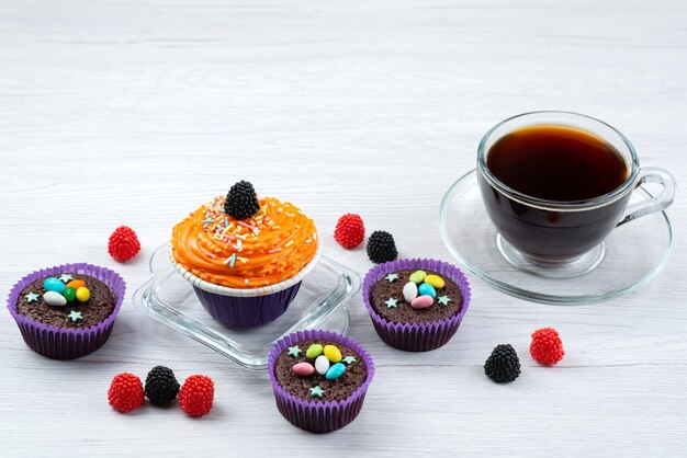 Una vista frontal de deliciosos brownies dentro de formas púrpuras junto con una taza de té en blanco, dulces de color caramelo