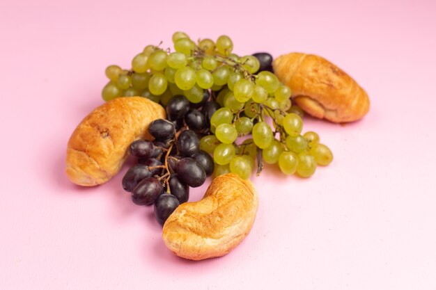 Foto gratuita vista frontal deliciosos croissants horneados con relleno de frutas junto con uvas negras y verdes frescas en el escritorio rosa