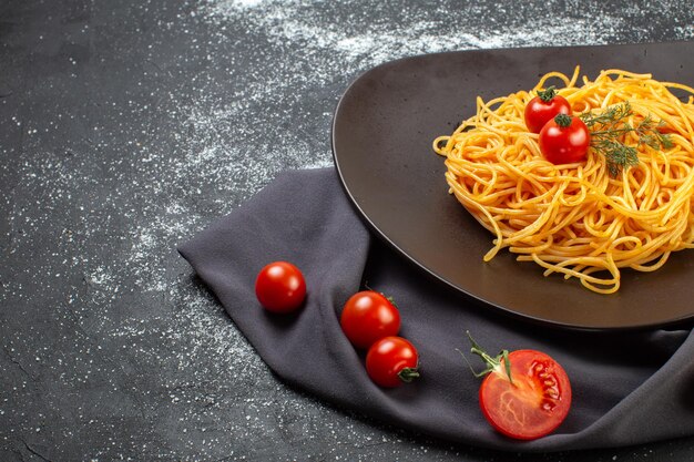 Vista frontal de deliciosos espaguetis en un plato negro sobre una toalla de color oscuro pastas crudas tomates frescos en el lado izquierdo sobre una mesa de color oscuro