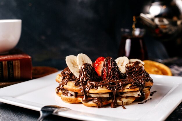 Vista frontal deliciosos panqueques de chocolate con plátanos frescos en rodajas y fresas dentro de un plato blanco sobre la superficie brillante