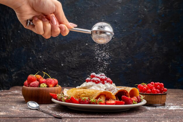 Una vista frontal deliciosos panqueques enrollados con arándanos y fresas rojas frescas obteniendo azúcar en polvo en el escritorio de madera comida baya de frutas