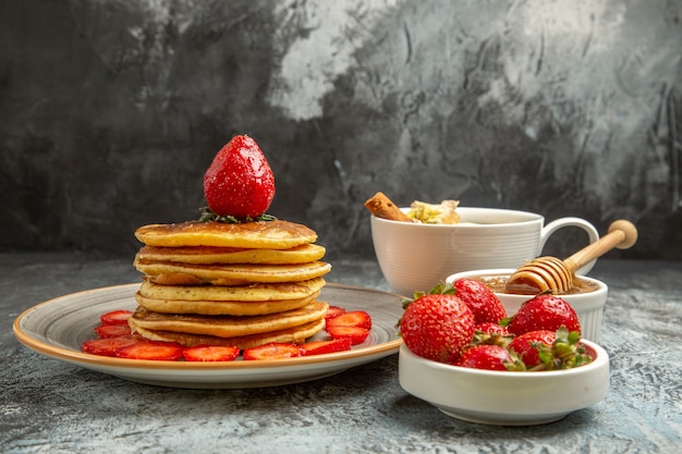 Vista frontal deliciosos panqueques con fresas y una taza de té sobre una superficie liviana pastel de frutas dulce