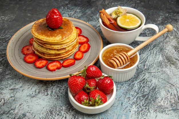 Vista frontal deliciosos panqueques con fresas y una taza de té en la superficie de la luz pastel de frutas dulces