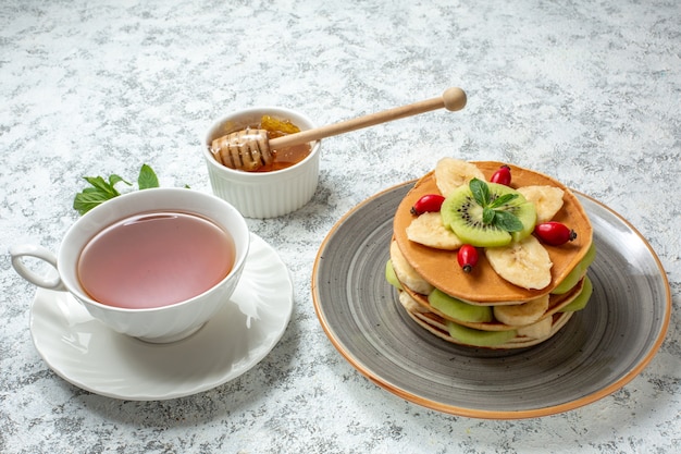 Vista frontal deliciosos panqueques con frutas en rodajas y una taza de té en la superficie blanca fruta postre dulce azúcar desayuno pastel de color