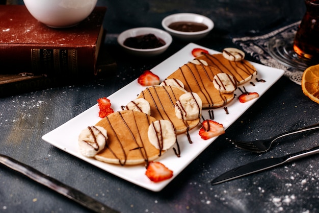 Foto gratuita vista frontal deliciosos panqueques junto con fresas rojas y plátanos dentro de un plato blanco sobre la superficie gris