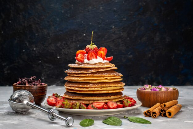 Una vista frontal deliciosos panqueques redondos con crema y fresas rojas en el pastel de escritorio ligero